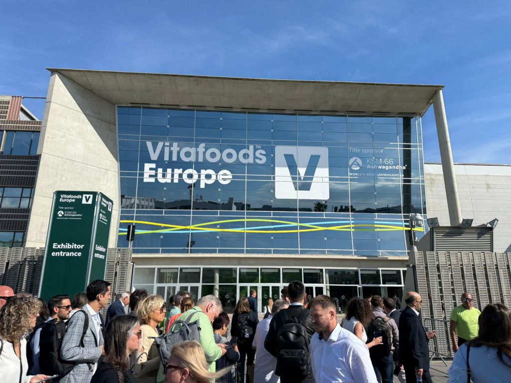 An image showing a crowd of business professionals in suits outside the Vitafoods Europe exhibition in Barcelona Spain. The picture shoes the Fira Barcelona Gran Via building which has Vitafoods Eutope and the logo on their glass window in white. The picture was taken by Arnie Ellison, Business Development Director at OPM (Labels & Packaging) Group.